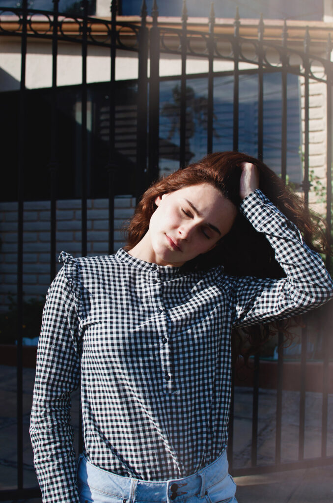 Sofia Renee a model with long brown hair is captured in a candid moment wearing a checkered shirt and blue jeans with her hand on her head standing in front of a house with a wrought iron gate.